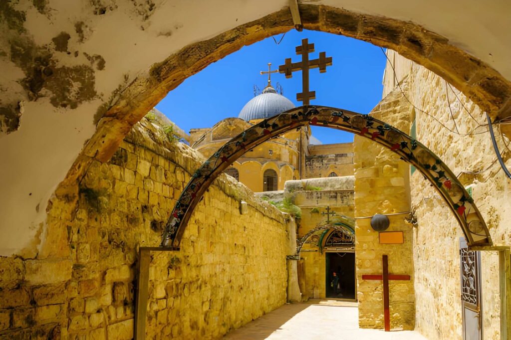 A perspective photograph taken from underneath a stone archway gate, looking directly into the courtyard of the Church of the Holy Sepulchre in Jerusalem. The image highlights the historic 12th-century Crusader facade with its carved stone arches. Above the facade, the large, dark grey dome of the church stands out against the sky, featuring its distinct open-top skylight oculus.