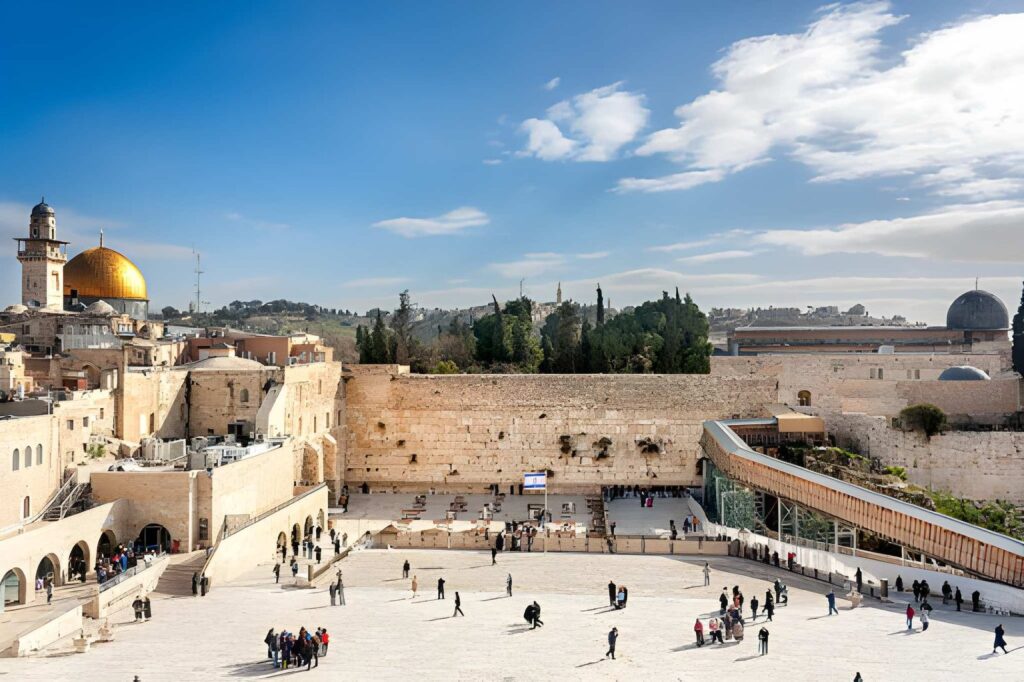 An aerial view of the Western Wall (Kotel) in Jerusalem captures the historic stone plaza filled with visitors alongside the expansive Al-Aqsa Mosque compound and the golden Dome of the Rock in the background.