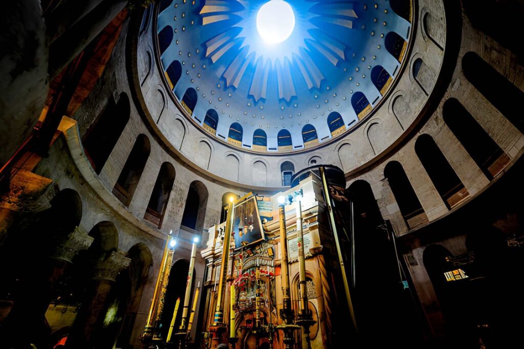 An interior photograph of the Aedicule, a small stone chapel shrine situated in the center of the massive church rotunda. The chapel doors or entrance are open, revealing the sacred tomb area inside, while natural sunlight streams down directly onto the structure from a large circular oculus skylight in the dome above.